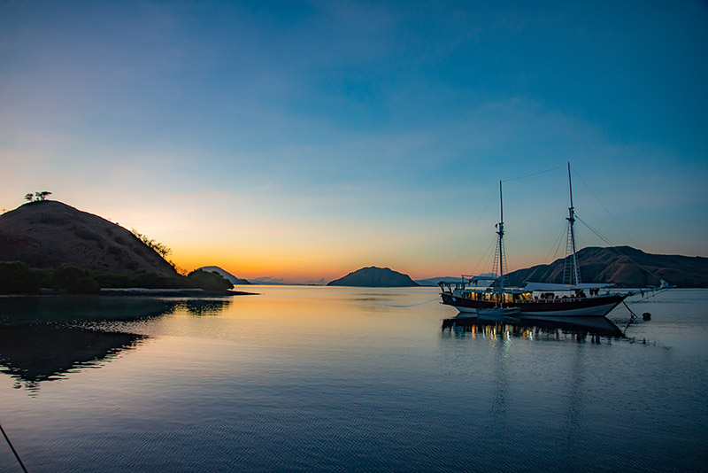 A two-masted boat at anchor in a still bay at dawn, hills on the horizon
