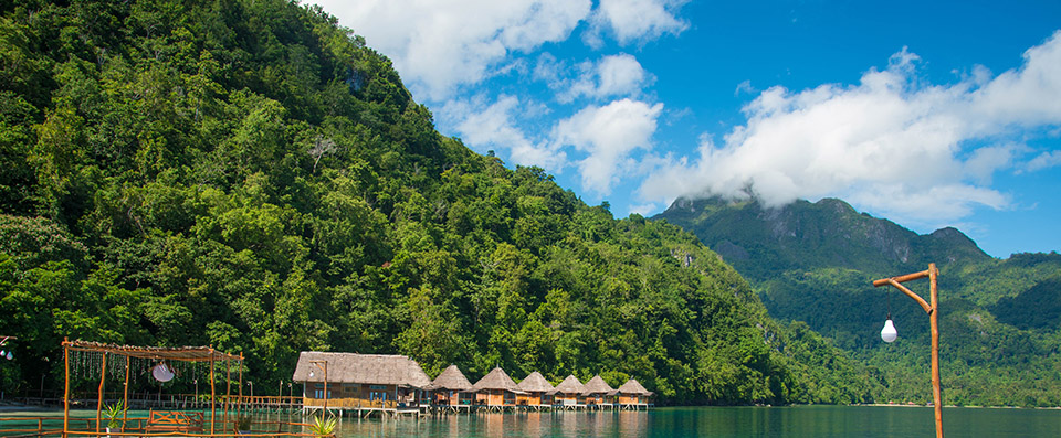 Thatched stilt cottages over calm water at the foot of a forested mountain, Indonesia